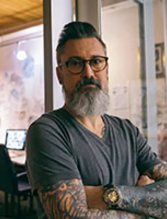 A thoughtful man with tattoos and glasses sitting in a cafe, possibly reflecting on the successful implementation of digital marketing strategies.