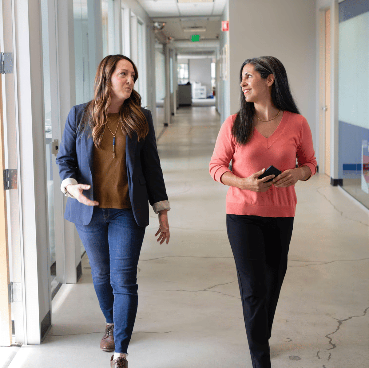 Two women walking and talking in a modern office hallway, one wearing a navy blazer and jeans, the other in a coral sweater holding a phone.