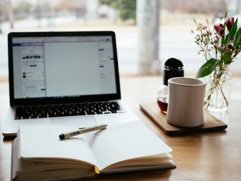 Open notebook with pen on a desk beside a laptop, coffee mug, and vase with flowers near a window.