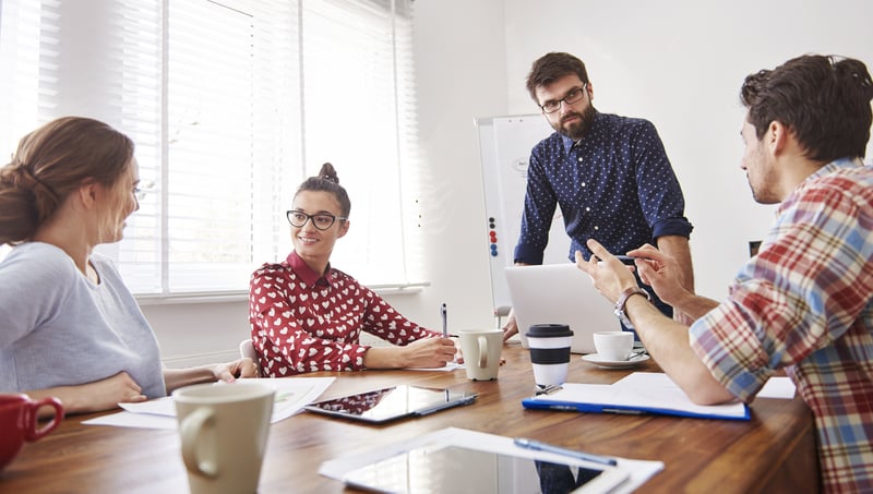 A diverse group of individuals gathered around a table, with laptops coffee cups and tablets discussing Project Management.