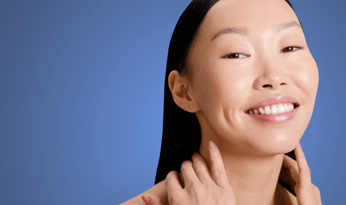 A woman with clear skin smiles while gently touching her neck. She has long, dark hair, and is set against a plain blue background.