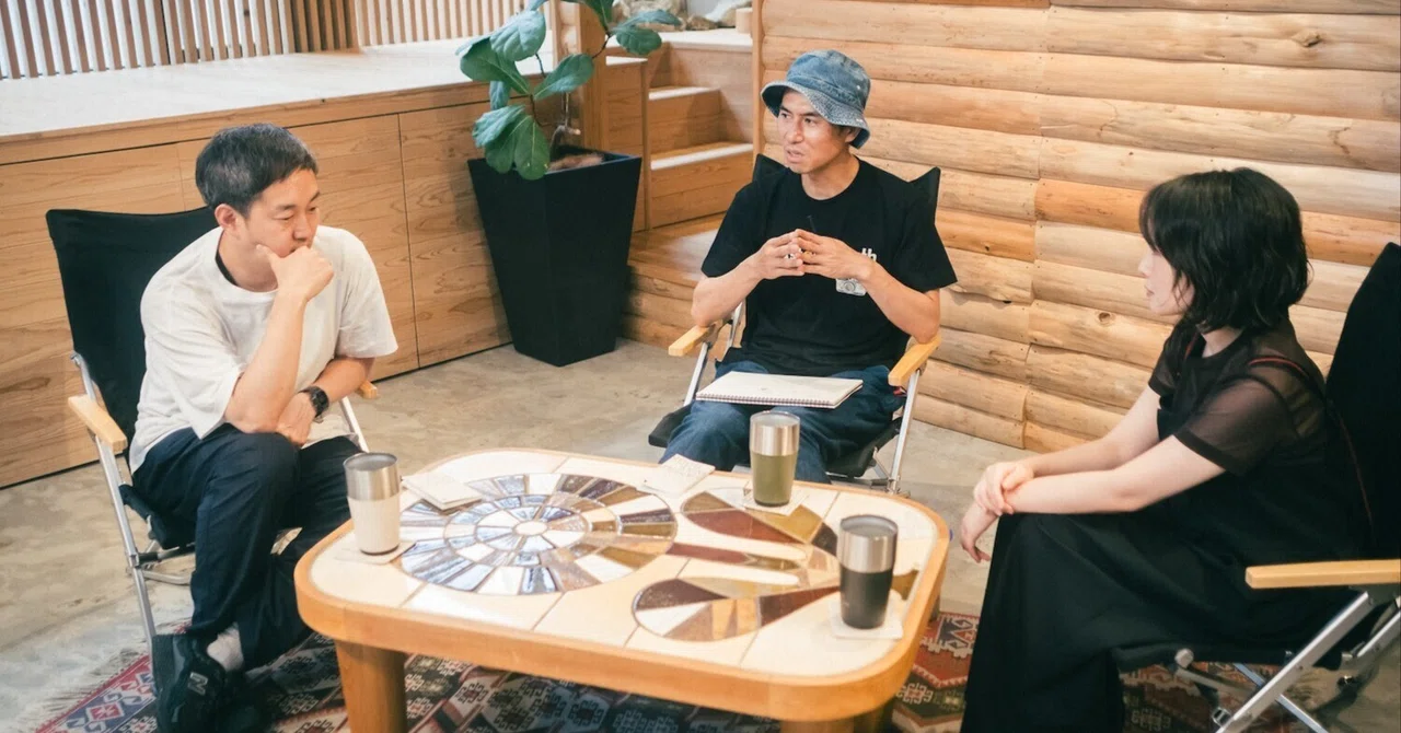 木の壁の部屋でテーブルを囲んで話す3人