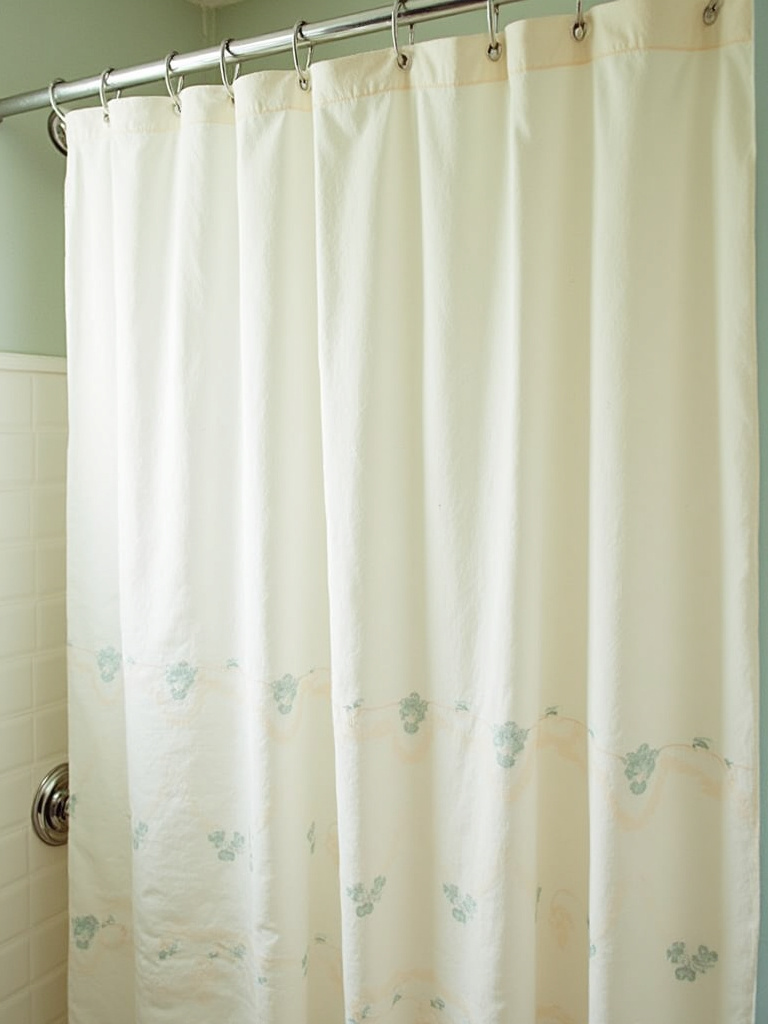 Detail of a vintage bathroom shower featuring a fabric shower curtain with a delicate floral pattern in soft pastel colors, with chrome shower fixtures visible and diffused lighting.