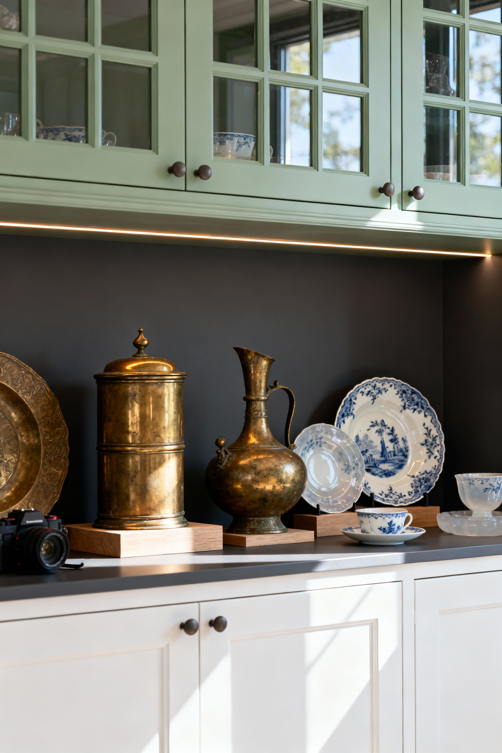 Traditional sage green kitchen cabinets with mullioned glass doors displaying a curated collection of heavy antique South Asian brass vessels and delicate blue-and-white fine china.