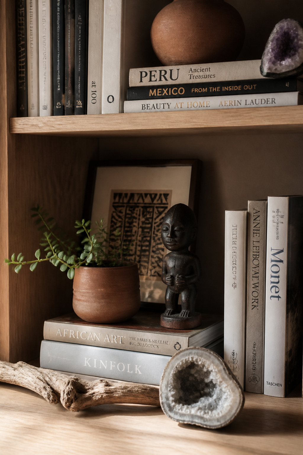 Eclectic bookshelf styling layers carved artifacts, terracotta vessels, horizontal book stacks, and negative space to create a composition that reads as genuinely collected rather than arranged.