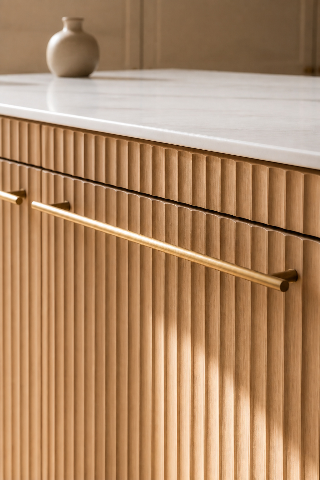 Fluted white oak cabinet panels cast shadow lines that deepen through the day as light shifts across the kitchen. The vertical grooves add tactile dimension that flat-panel cabinets simply cannot deliver.
