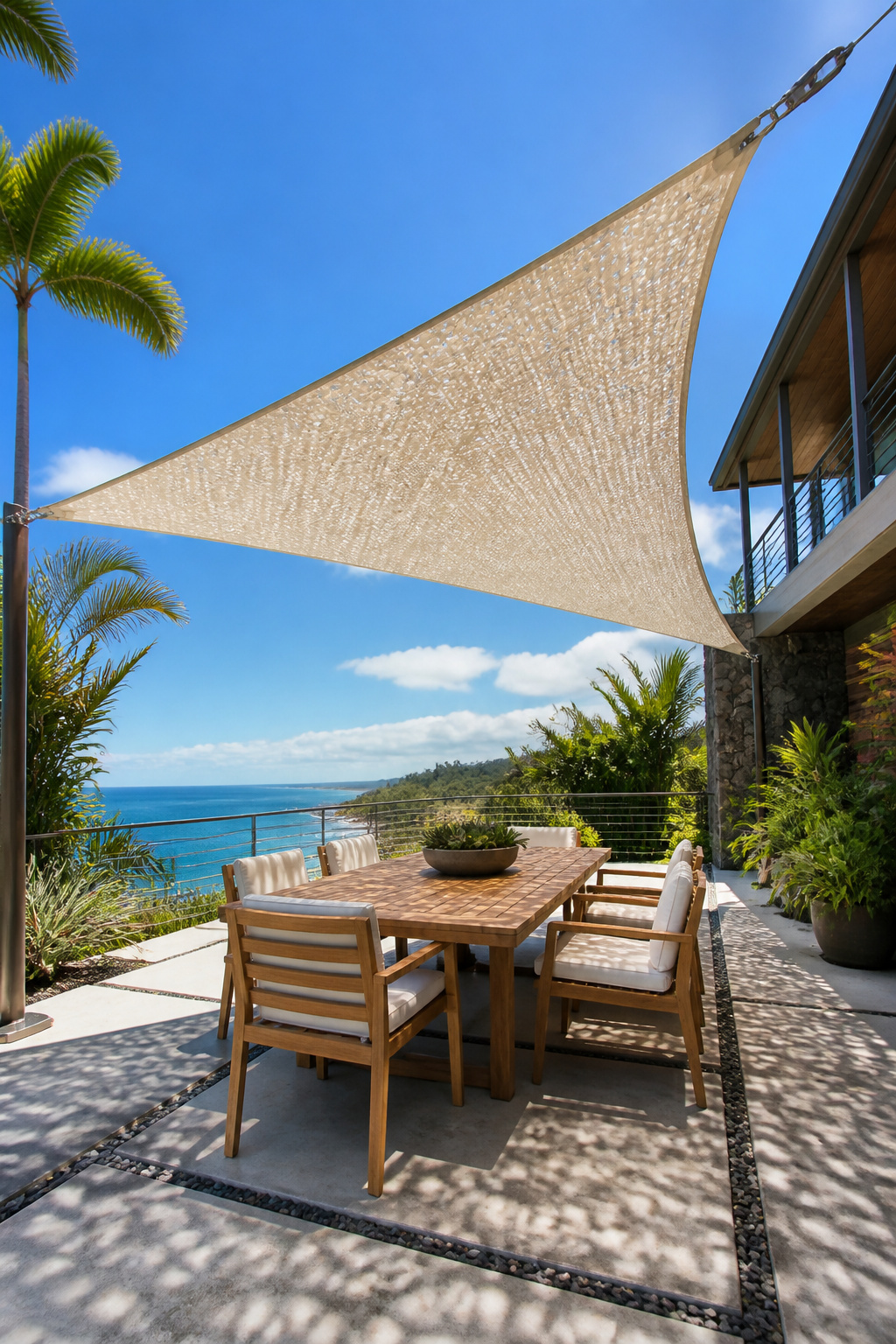 A triangular HDPE sail canopy in warm sand-white filters 95% of UV through its knitted mesh above a teak dining set, creating dappled light patterns on a coastal contemporary concrete paver patio.