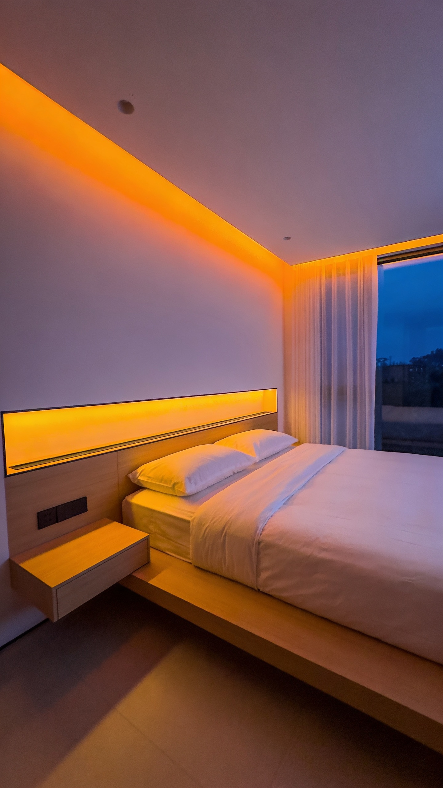 A warm amber-lit minimalist bedroom showcasing circadian smart lighting technology integrated into the wall behind a low platform bed.