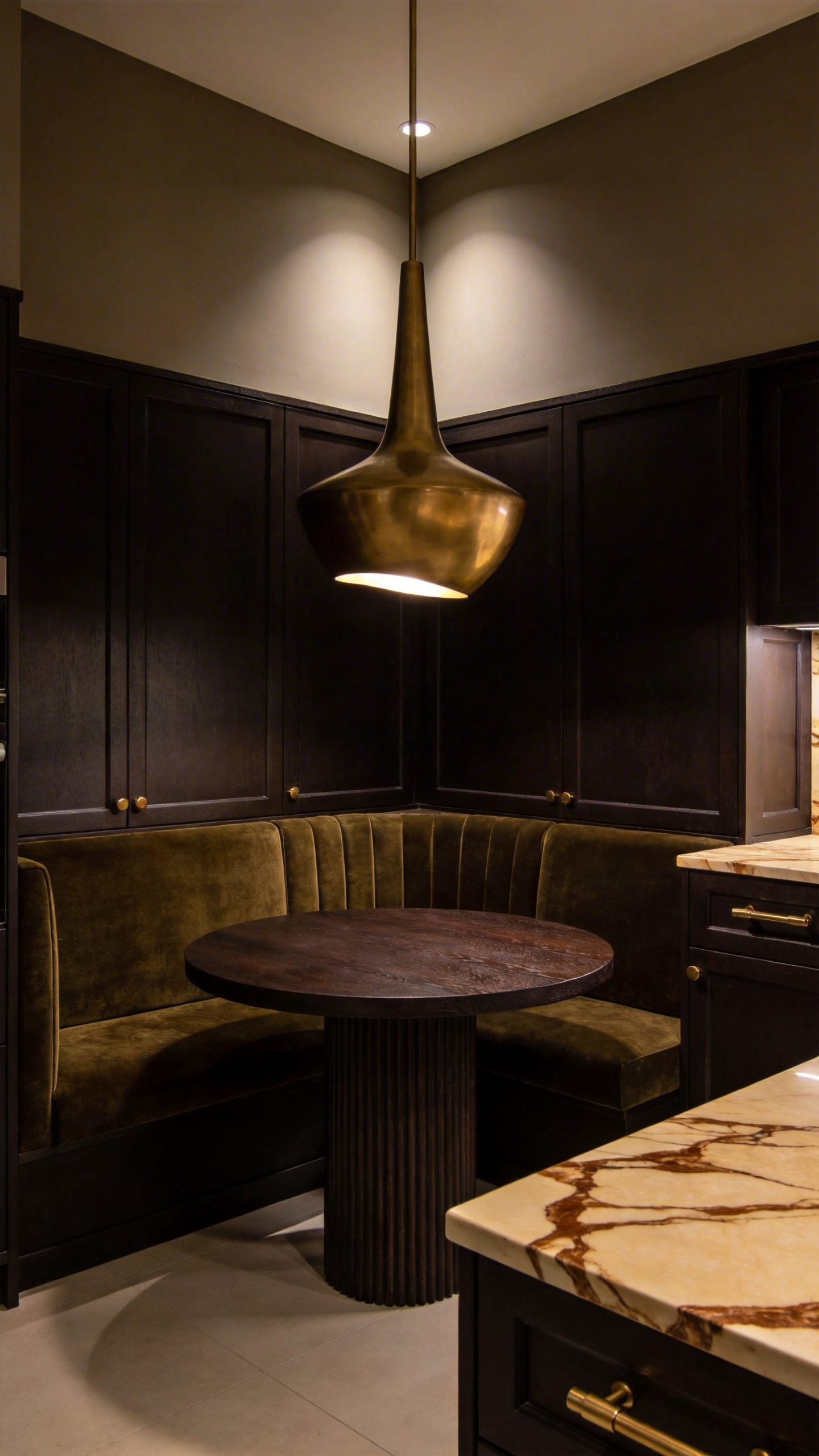 Dramatic and luxurious built-in dining nook featuring matte espresso brown cabinetry, olive velvet banquette seating, and a sculptural brass pendant light, conveying a moody, intimate atmosphere.