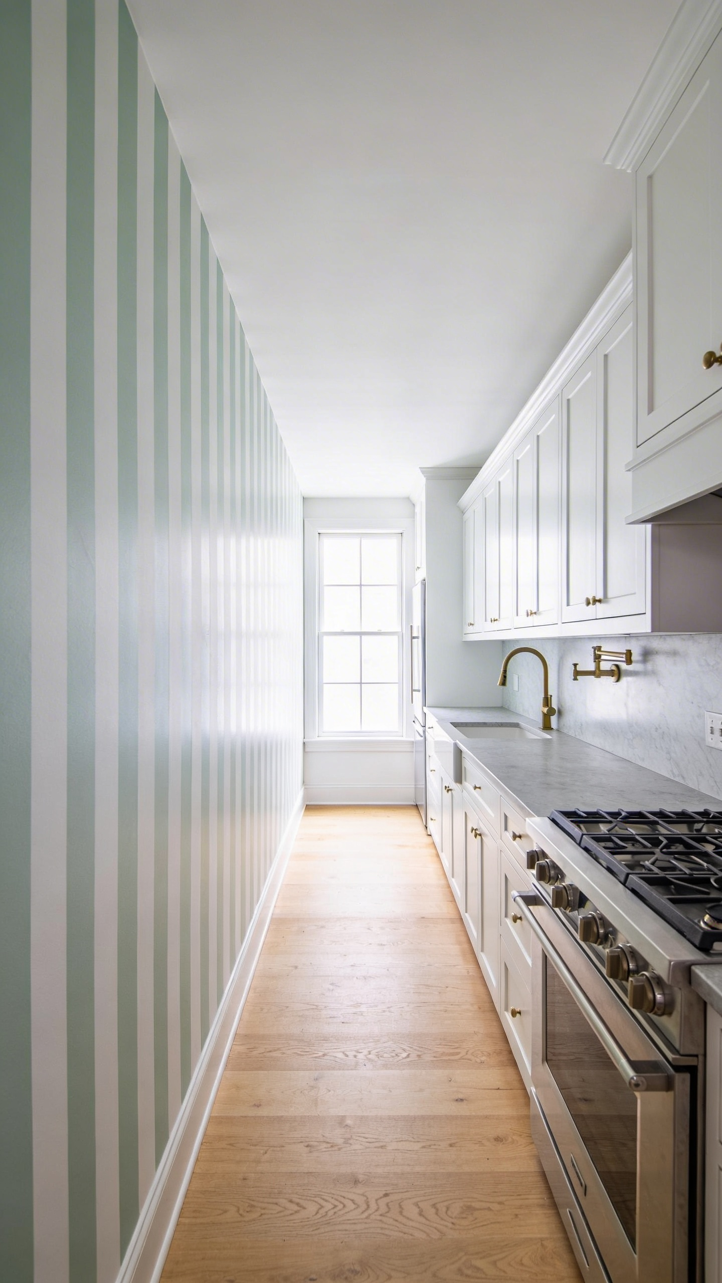 Narrow galley kitchen featuring white cabinets and vertically striped wallpaper in sage green and white to create the illusion of higher ceilings and increased space.
