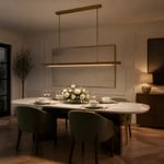 An aged brass linear suspension at the correct 33-inch height above a marble dining table — the proportions, the colour temperature, and the supplemental lighting all working together in a room that's ready for the evening.