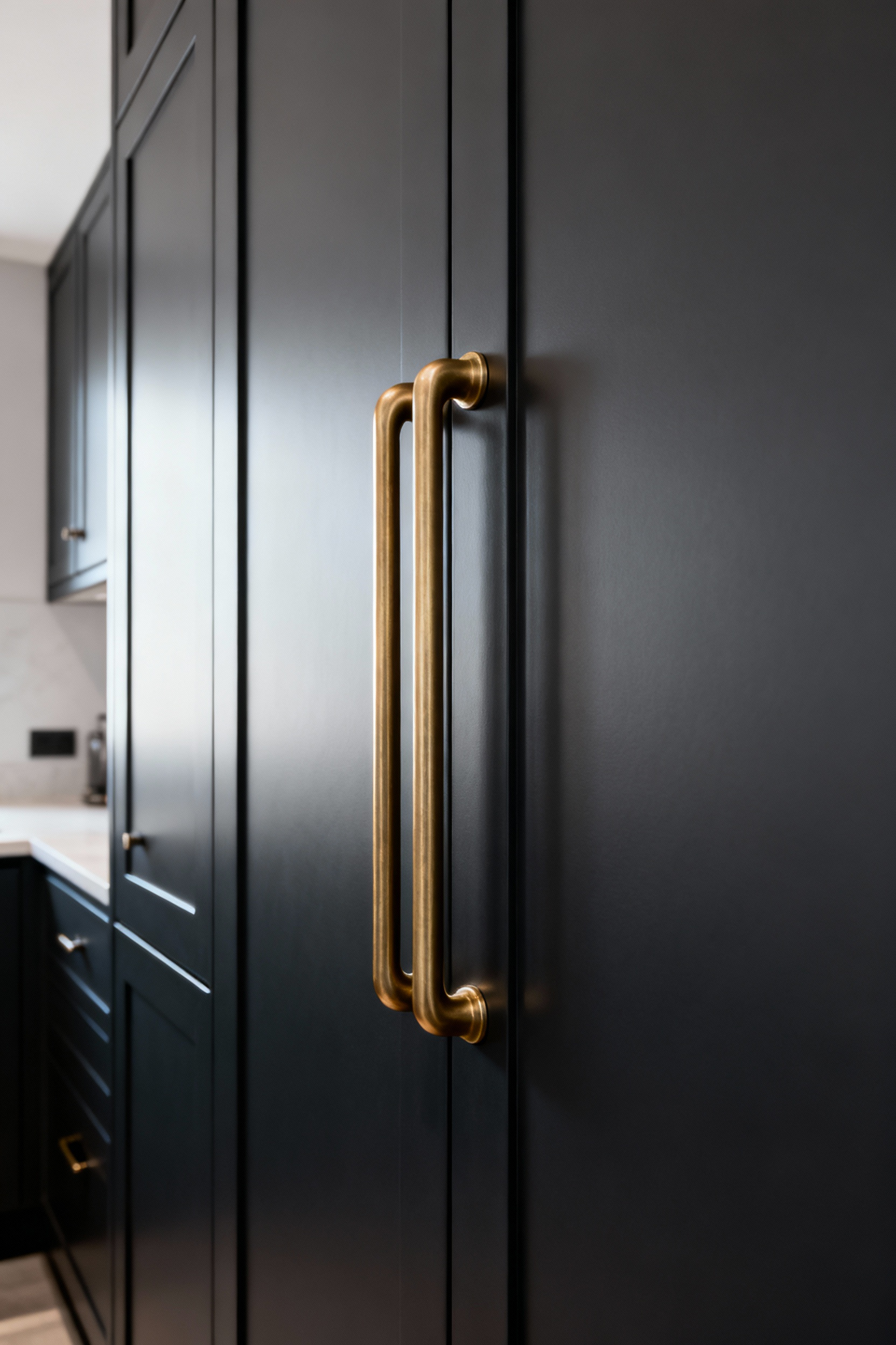 A tall, dark kitchen pantry cabinet featuring a long, vertical brass pull handle, designed for ergonomic comfort and easy reach on a substantial door.