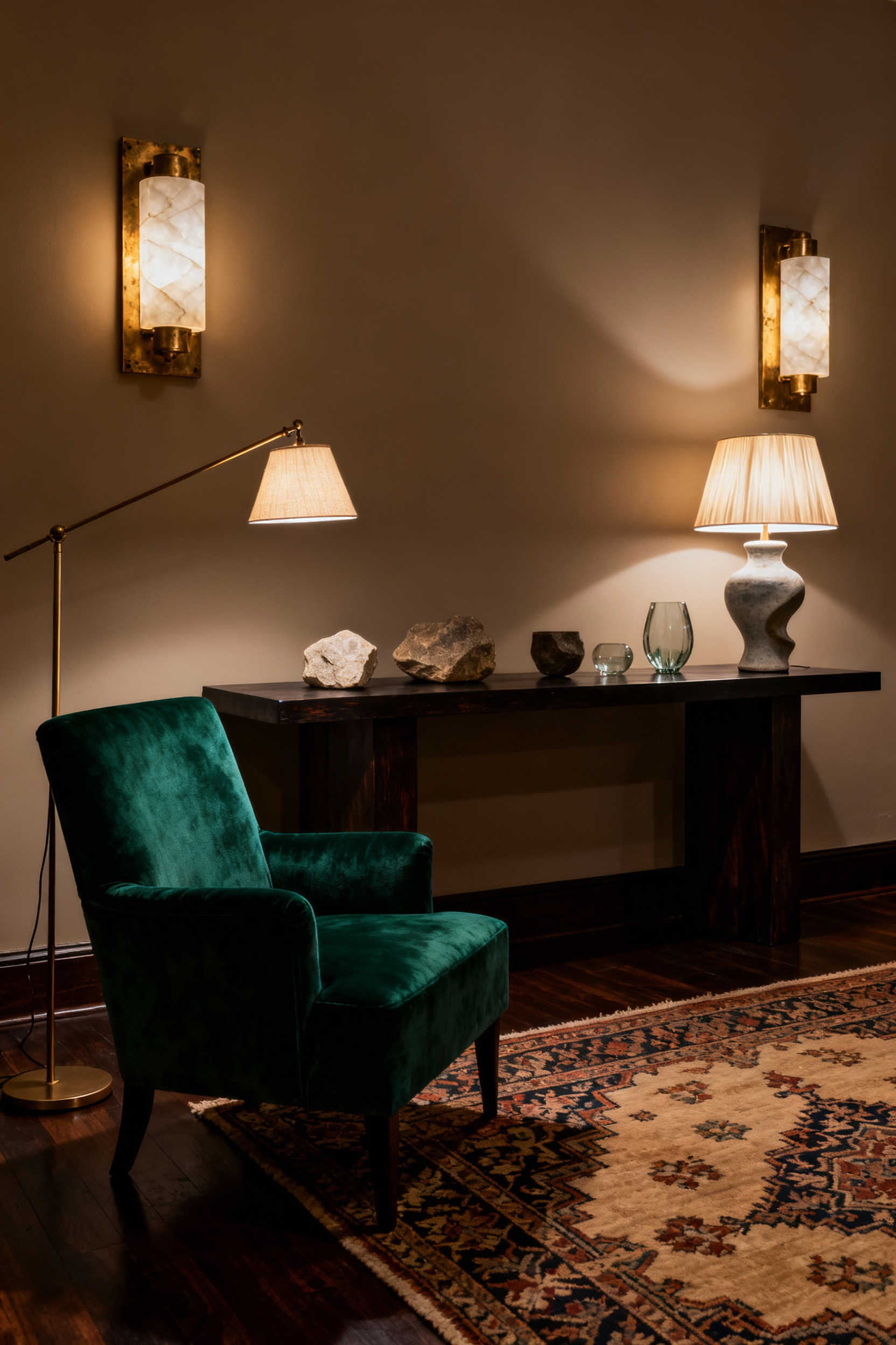 A luxurious, transitional living room in the evening, showcasing layered lighting using dimmable alabaster wall sconces, a brass floor lamp, and a table lamp, creating a warm, atmospheric glow against an emerald velvet sofa and dark wood paneling.