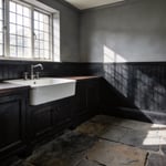 A moody and sophisticated laundry room featuring deep charcoal beadboard walls, a farmhouse sink, and slate floors inspired by historical Victorian scullery designs.