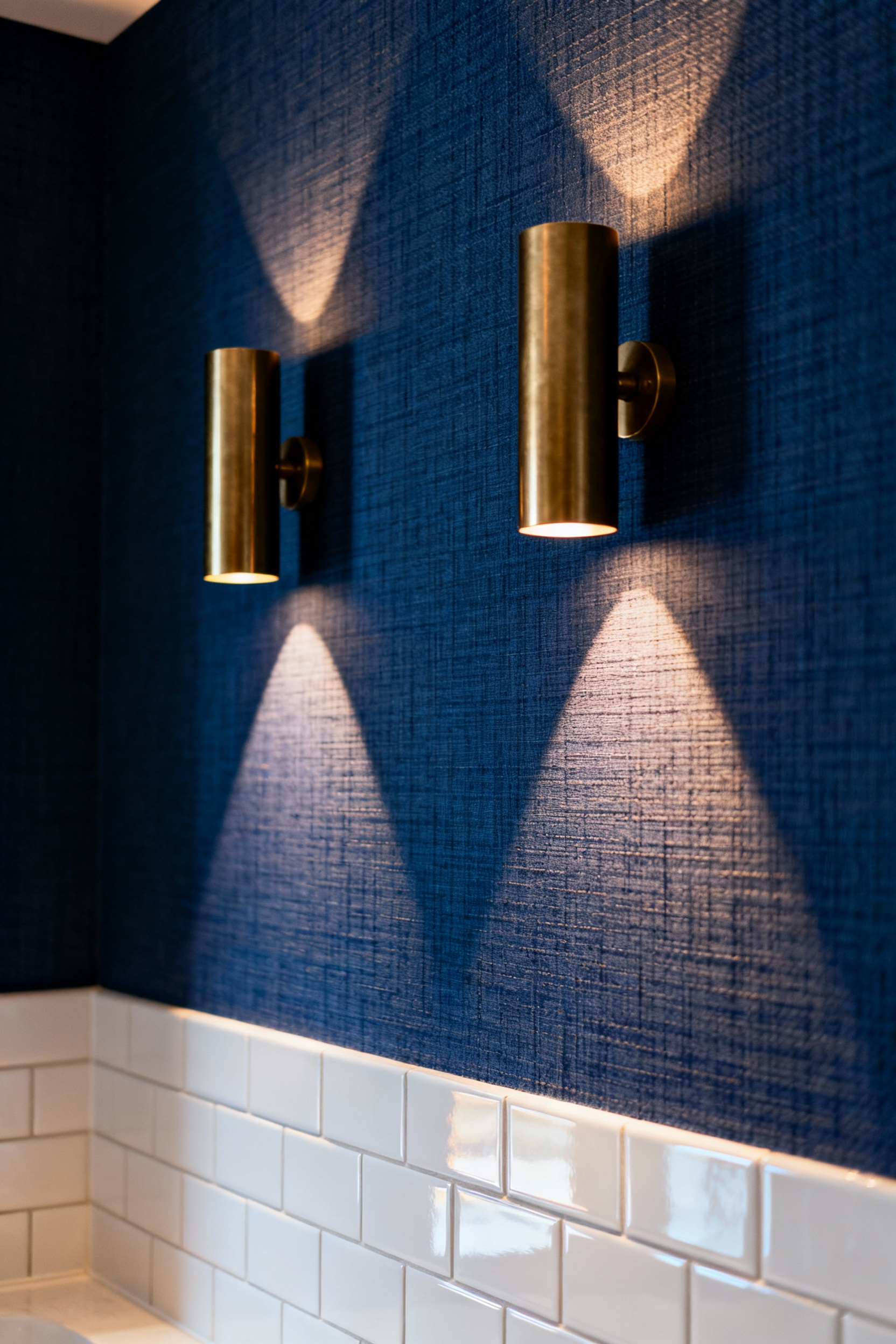 A contemporary bathroom wall featuring deep indigo woven grasscloth wallpaper dramatically lit by two brushed brass cylindrical sconces mounted at eye level, emphasizing the textured shadow play above a glossy white tiled wainscoting.