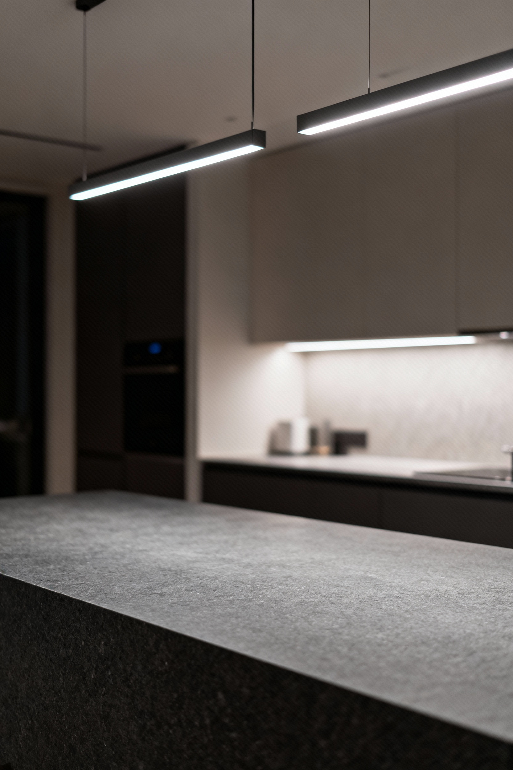 Honed charcoal-grey quartz kitchen island countertop showcasing diffuse light from linear LED pendants, highlighting texture and depth without glare, in a modern kitchen setting.