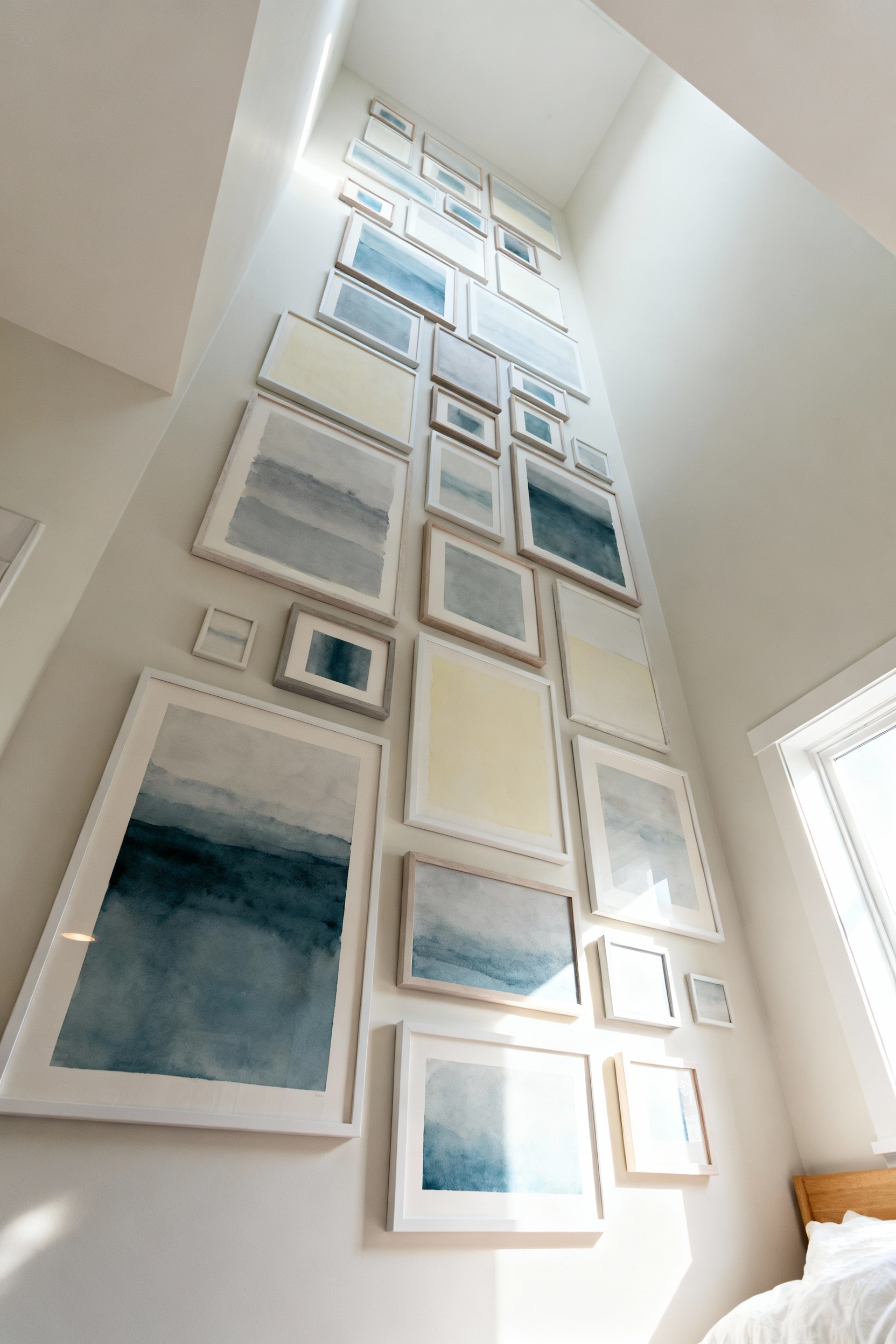 Vertical gallery wall in a serene small bedroom, featuring varied framed abstract art extending high towards the ceiling, enhancing perceived room height. Soft, natural light illuminates the display, showing a clean, minimalist aesthetic.