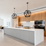 A wide view of a minimalist open-concept modern kitchen featuring a large white quartz island, light gray handleless cabinets, and light oak wood storage, designed to accommodate multiple users and collaboration.