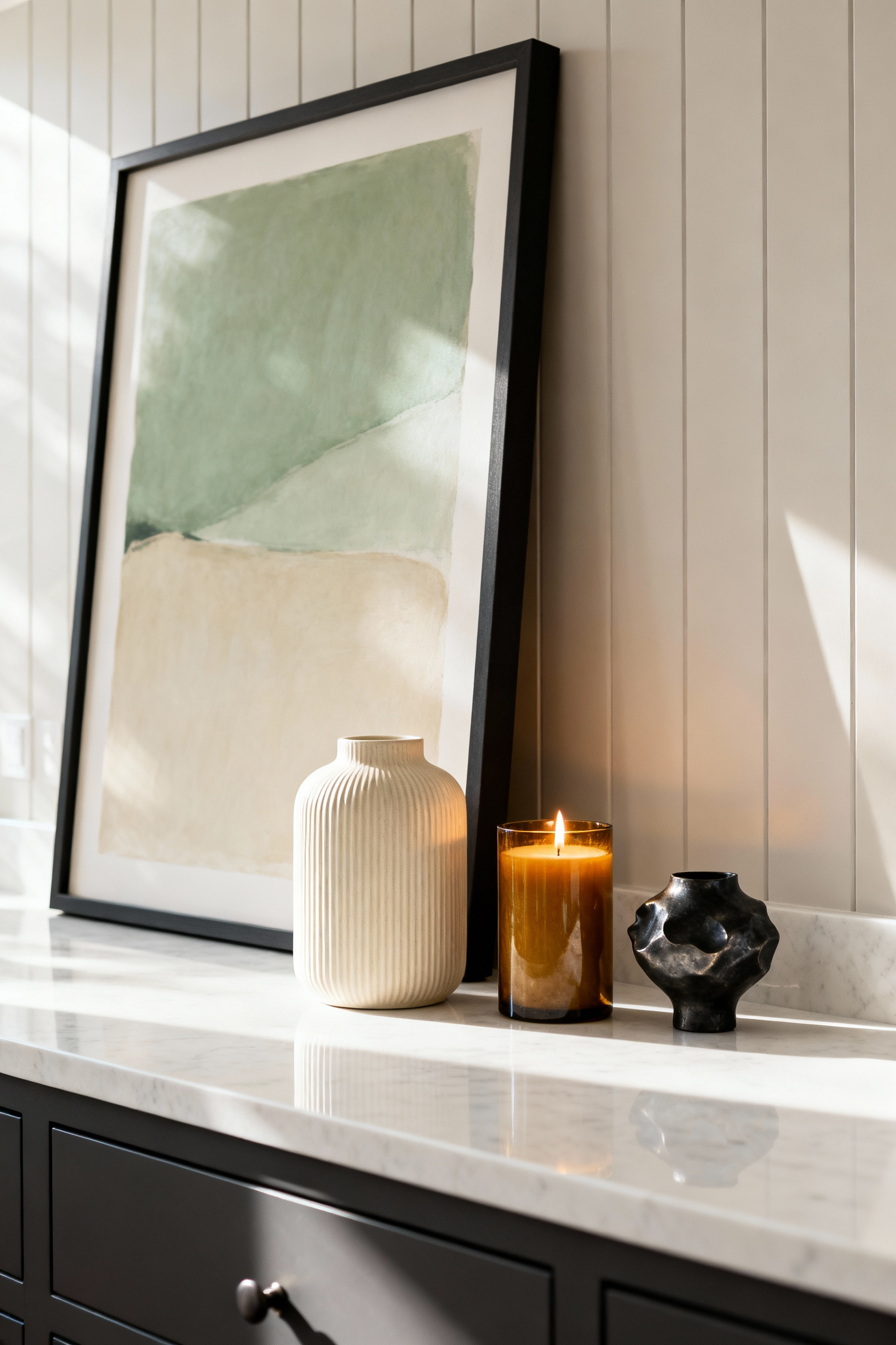 A sophisticated bathroom vanity counter displaying the 'Lean' art technique, featuring a large framed abstract piece anchored against the wall with smaller, layered decor items like a lit candle and ribbed ceramic vase to maximize visual depth.