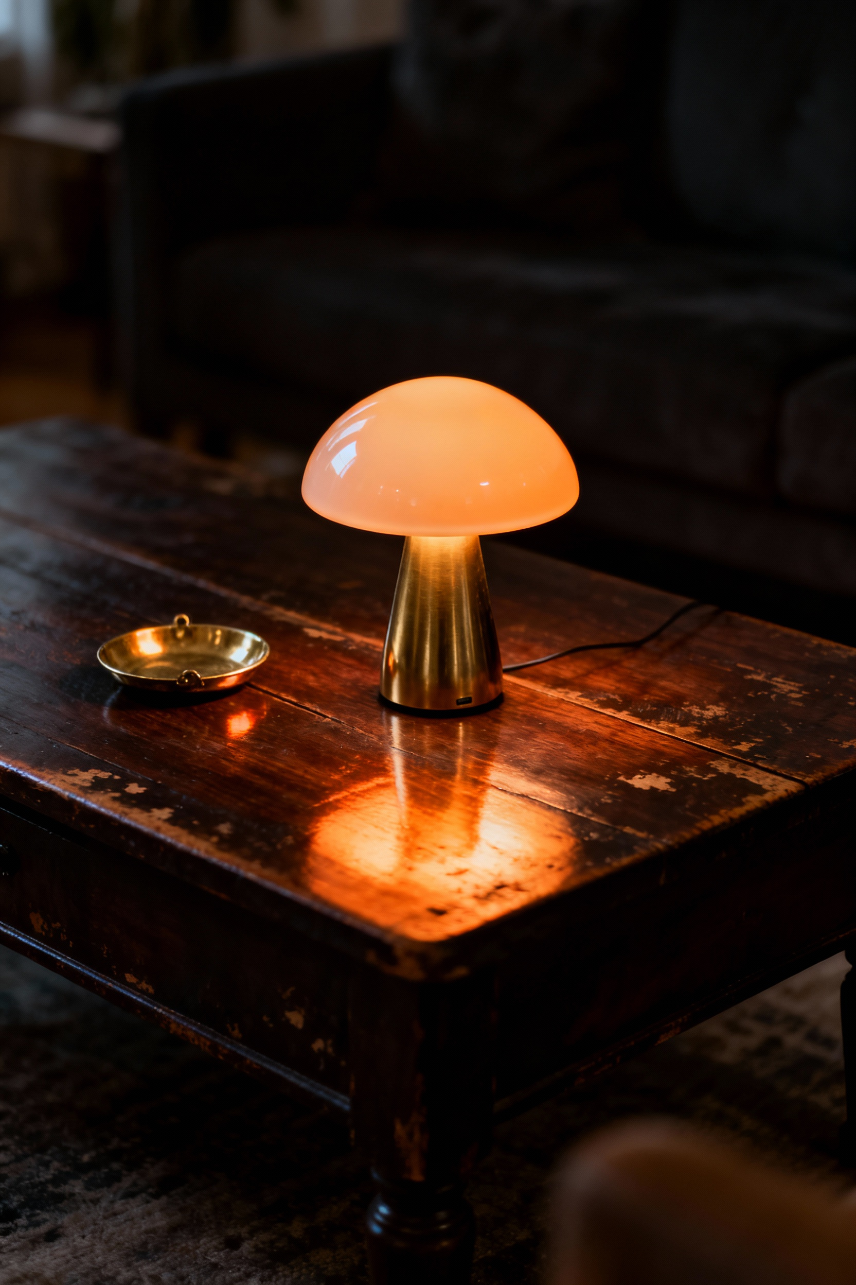 A close-up of a vintage walnut coffee table illuminated solely by the ultra-warm, amber light (2200K) cast by a sculptural brass and opal glass mushroom lamp.