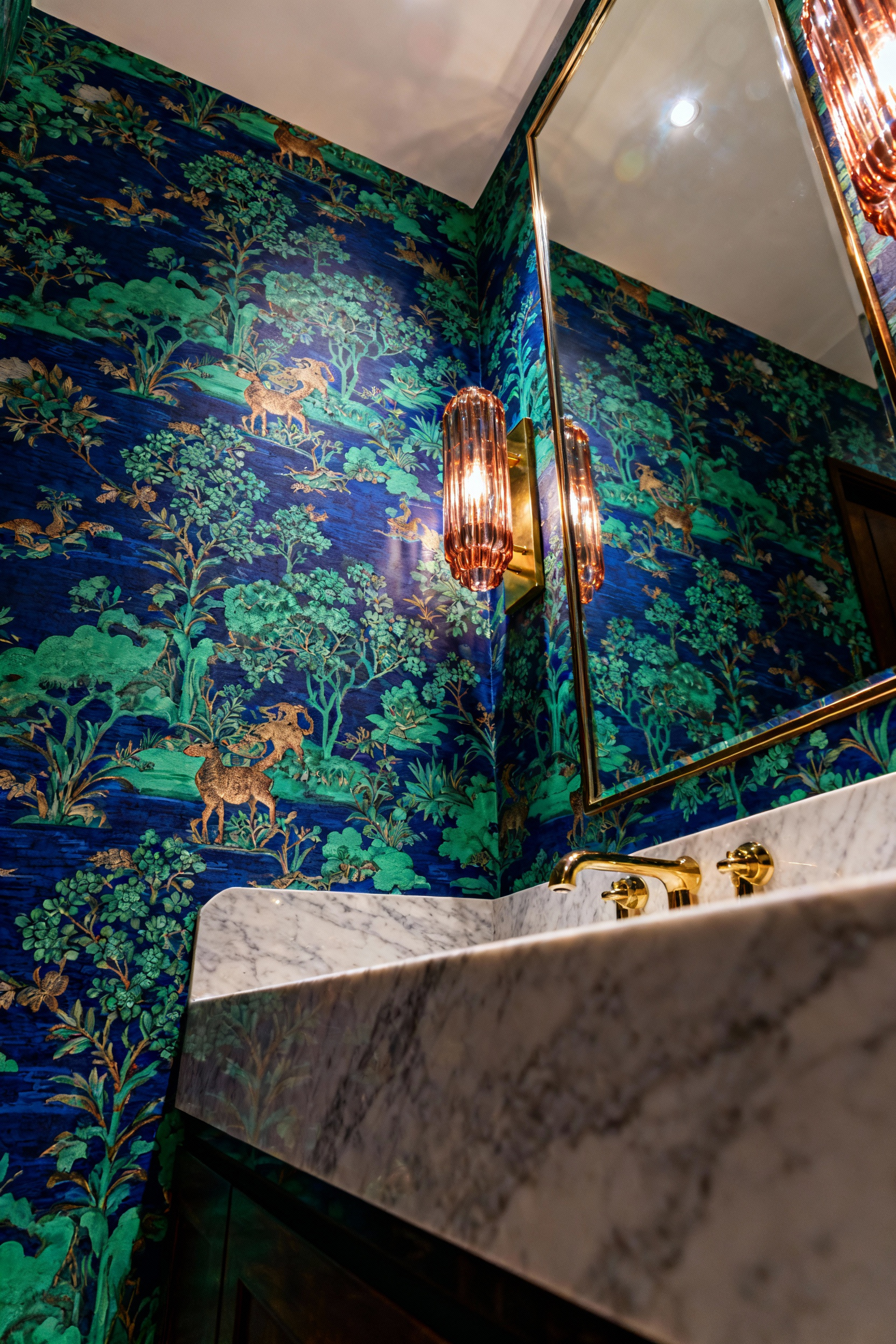 A maximalist bathroom accent wall featuring luxurious, intricate Chinoiserie wallpaper with symbolic flora and fauna, evoking personal mythology. Soft lighting illuminates the rich blue and emerald tones with gold metallic accents.