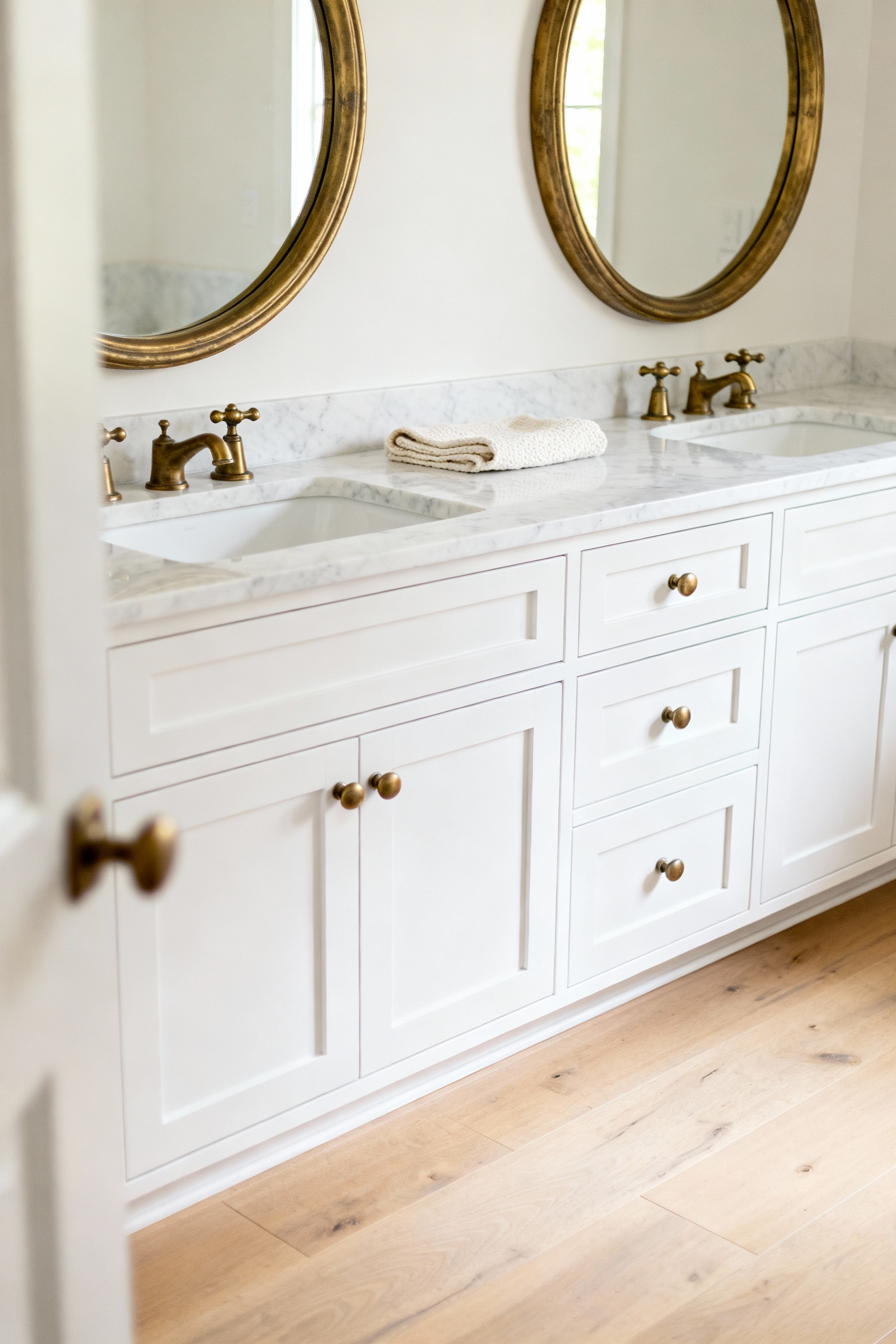 A modern, double-sink white bathroom vanity featuring unlacquered brass hardware and a marble countertop, set against natural wood flooring, demonstrating how warm metals counteract cool white surfaces.