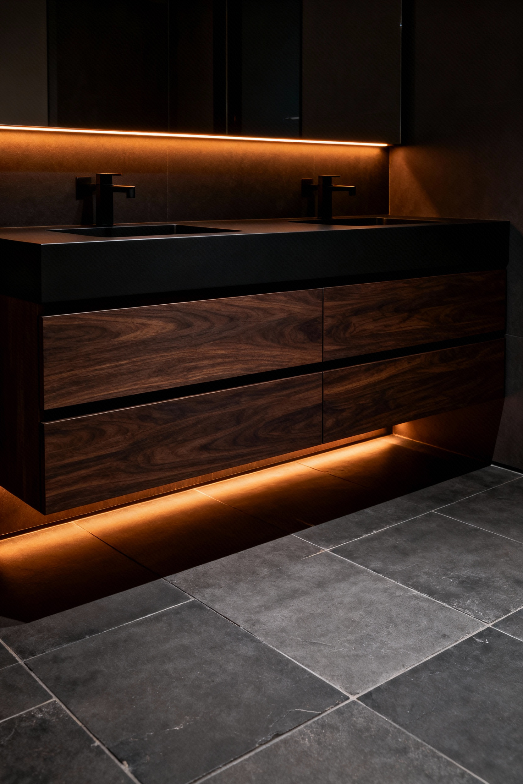 A modern floating dark wood bathroom vanity illuminated solely by warm amber toe-kick LED night lighting, casting a low glow across the dark slate floor for safe navigation.