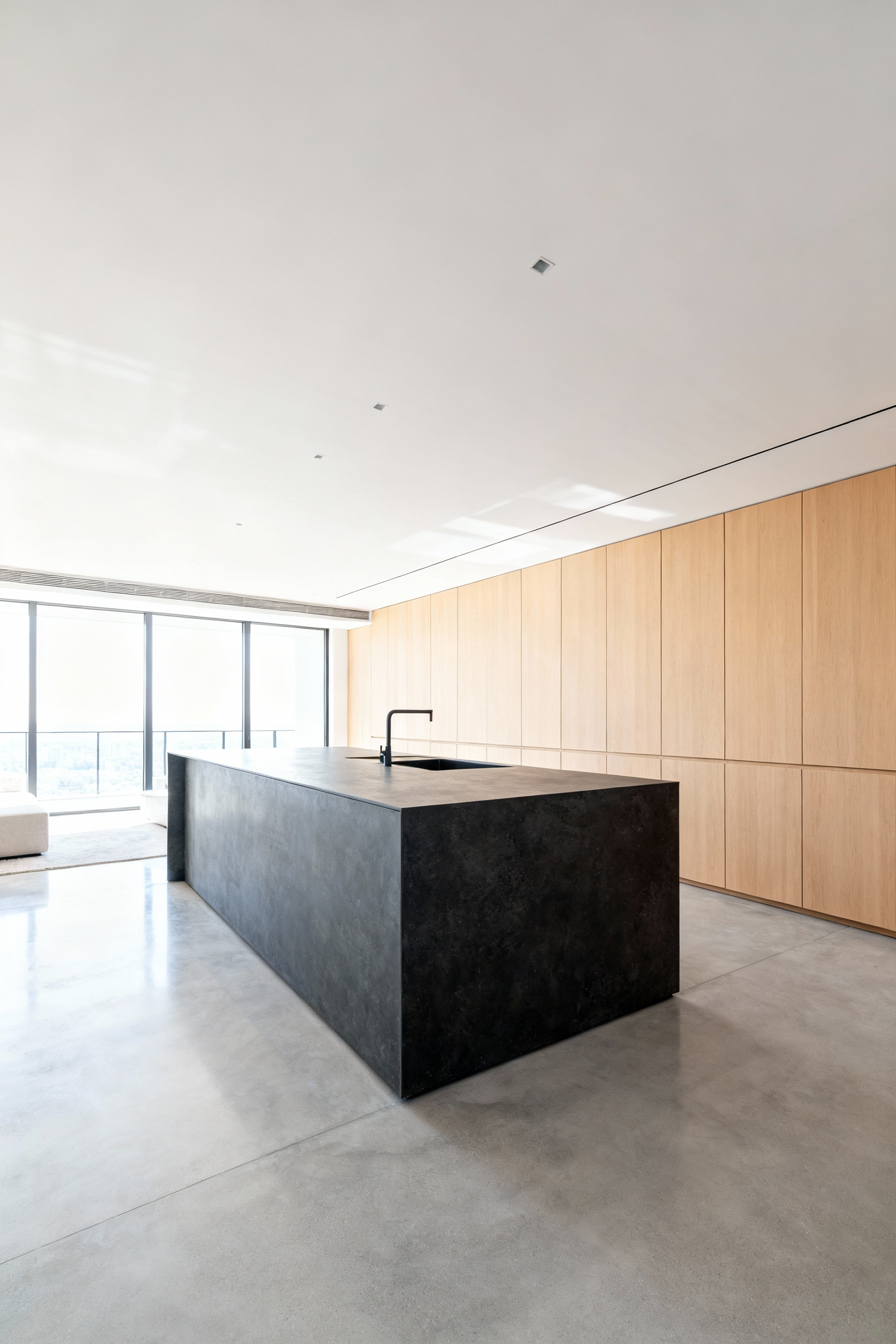 A spacious modern open-plan kitchen featuring a large monolithic dark stone island acting as the central social and architectural anchor, illuminated by bright natural light.