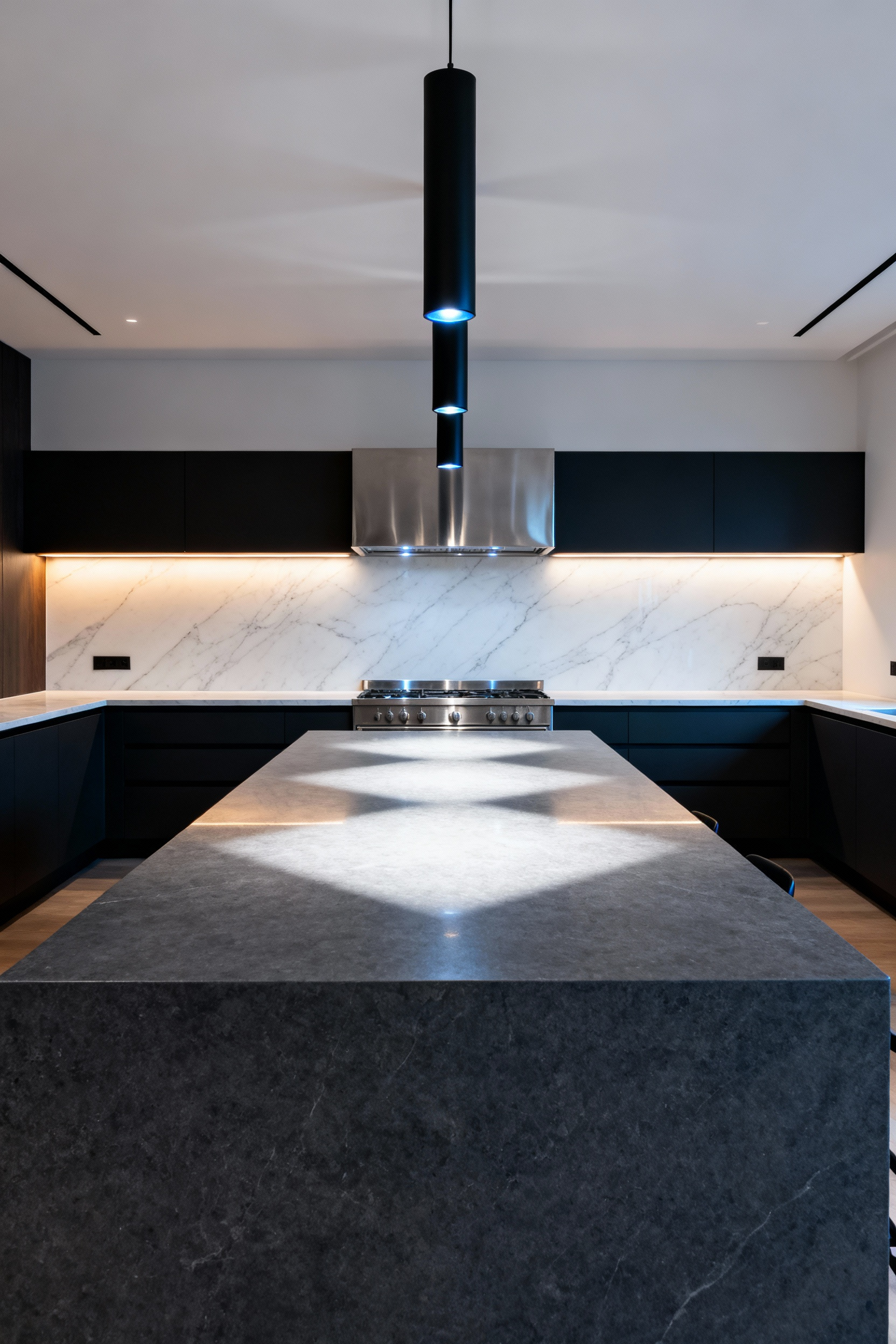 A wide view of a modern kitchen showing three black cylinder pendant lights centered directly beneath a dominant stainless steel range hood, balancing the sightline above a large gray kitchen island.