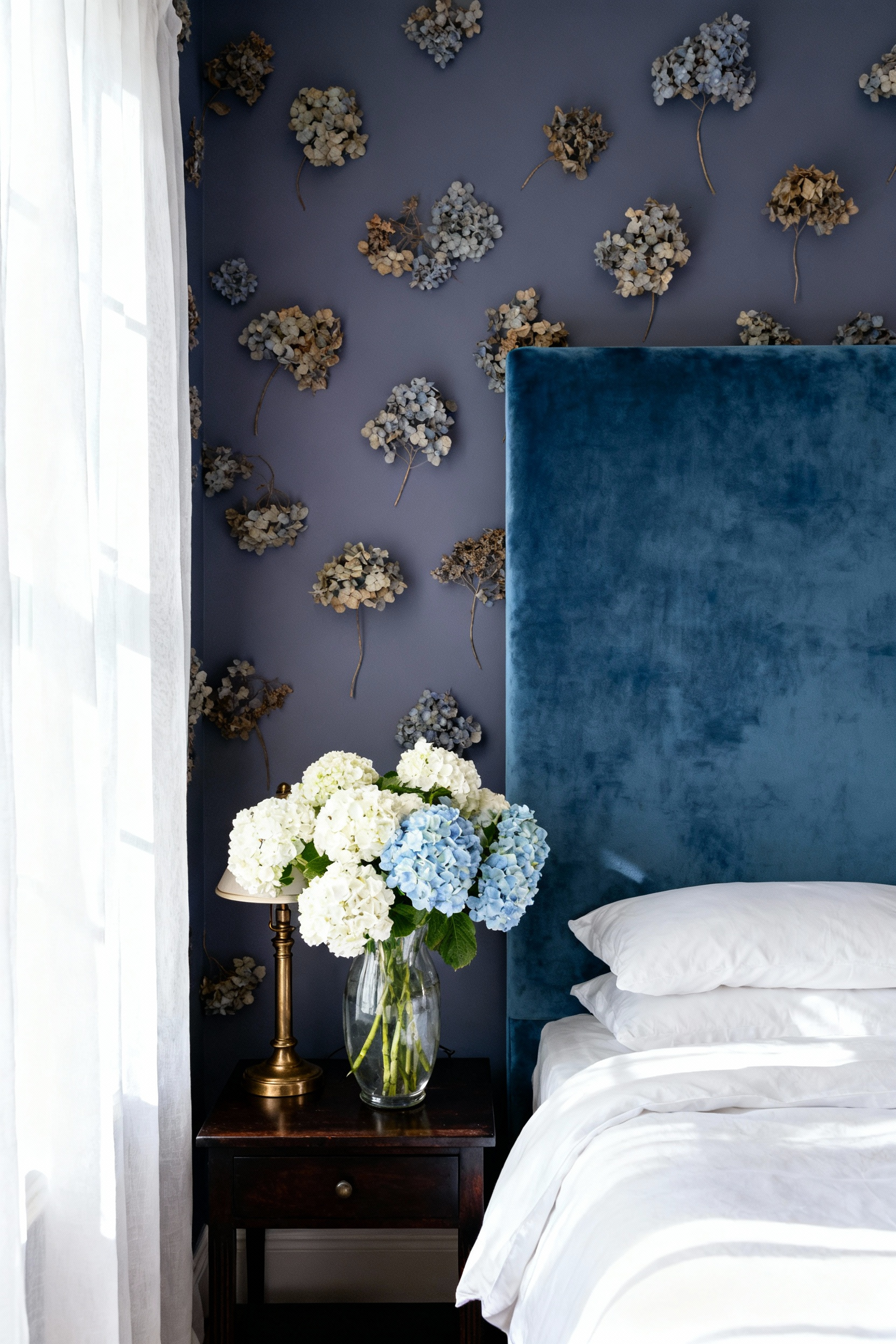 A tranquil bedroom featuring walls and an upholstered headboard painted in a sophisticated grayish-purple hydrangea blue, complemented by white linen bedding and natural daylight.