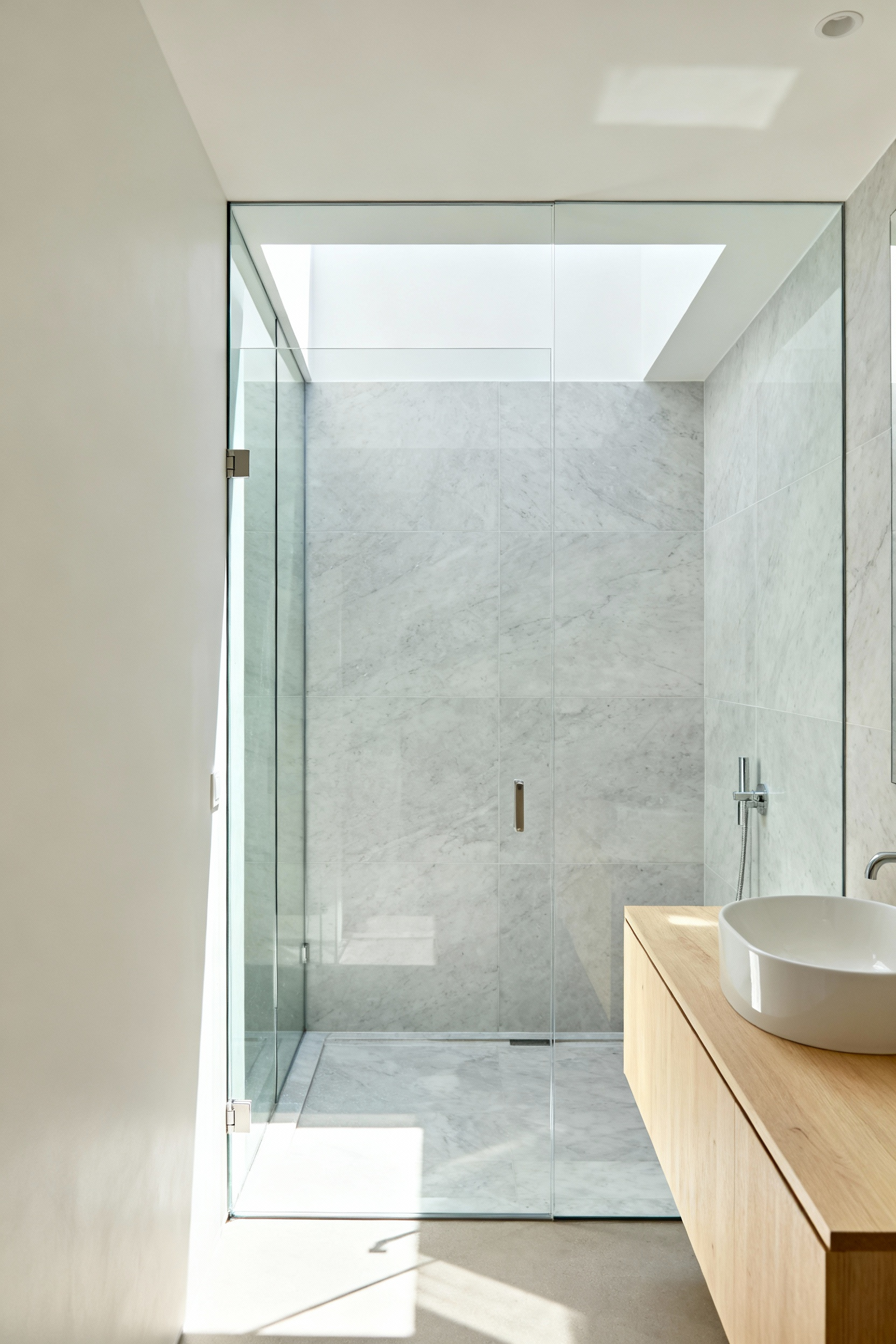 A small modern bathroom featuring a completely frameless, transparent glass shower partition designed to eliminate visual barriers and extend sightlines, creating an illusion of a much larger room.