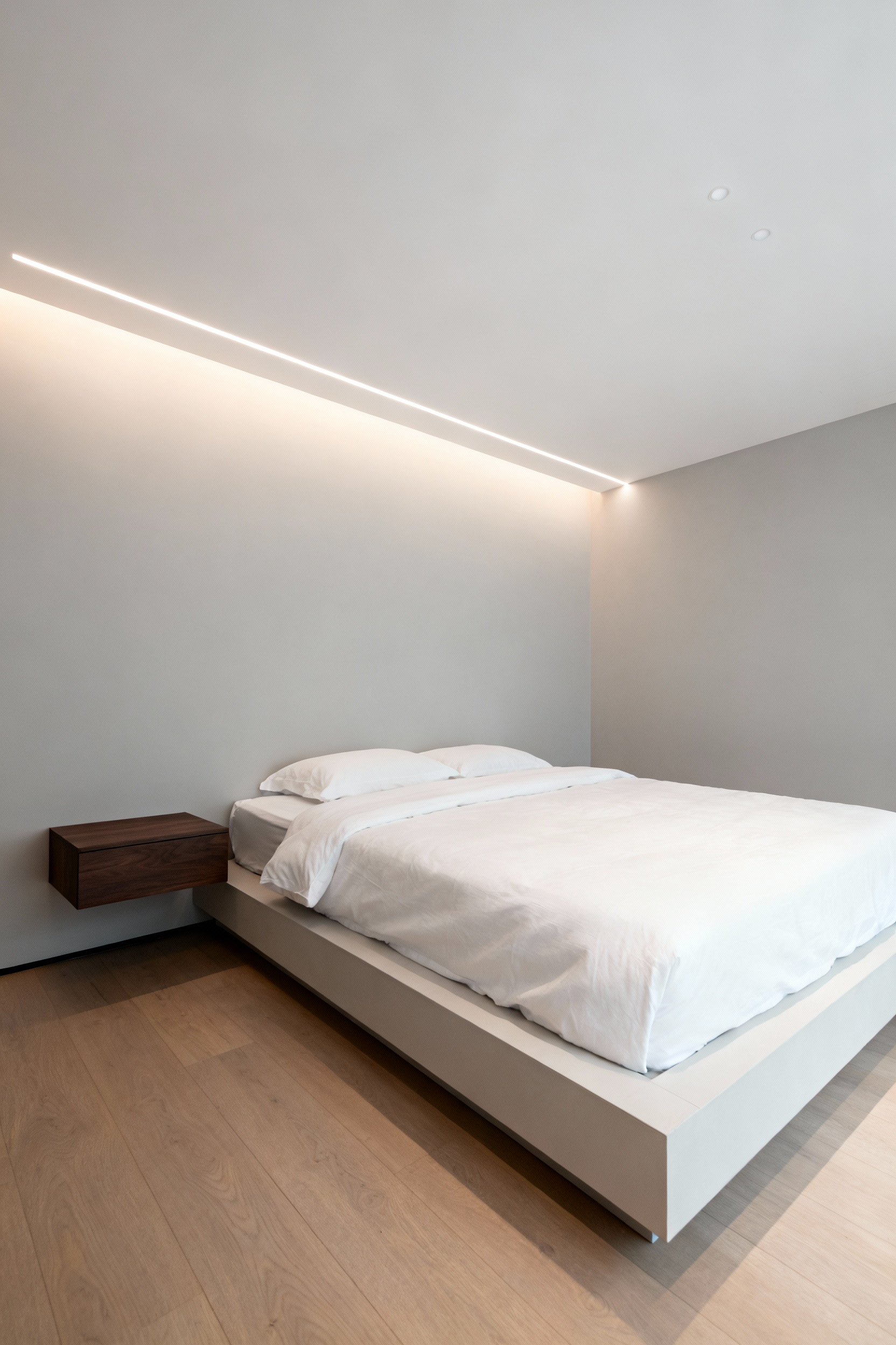 A wide view of a modern minimalist bedroom featuring a matte white ceiling with integrated trimless recessed beam lighting that provides soft, warm illumination over a low-profile platform bed.