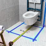 A photograph of a small, renovated bathroom with light gray tile showing measuring tape and blue lines on the floor indicating the precise 30-inch clearance zone required for a toilet fixture according to building codes.