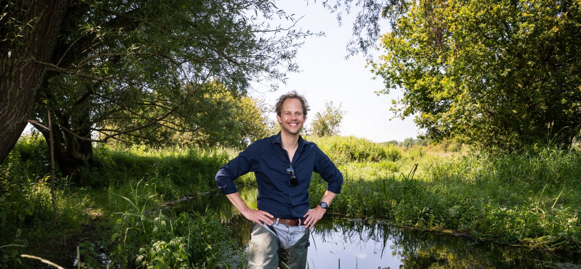 Man die tot net onder zijn middel in het water staat, hij heeft lieslaarsen aan en kijkt lachend in de camera