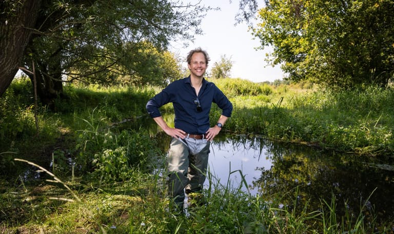 Man die tot net onder zijn middel in het water staat, hij heeft lieslaarsen aan en kijkt lachend in de camera
