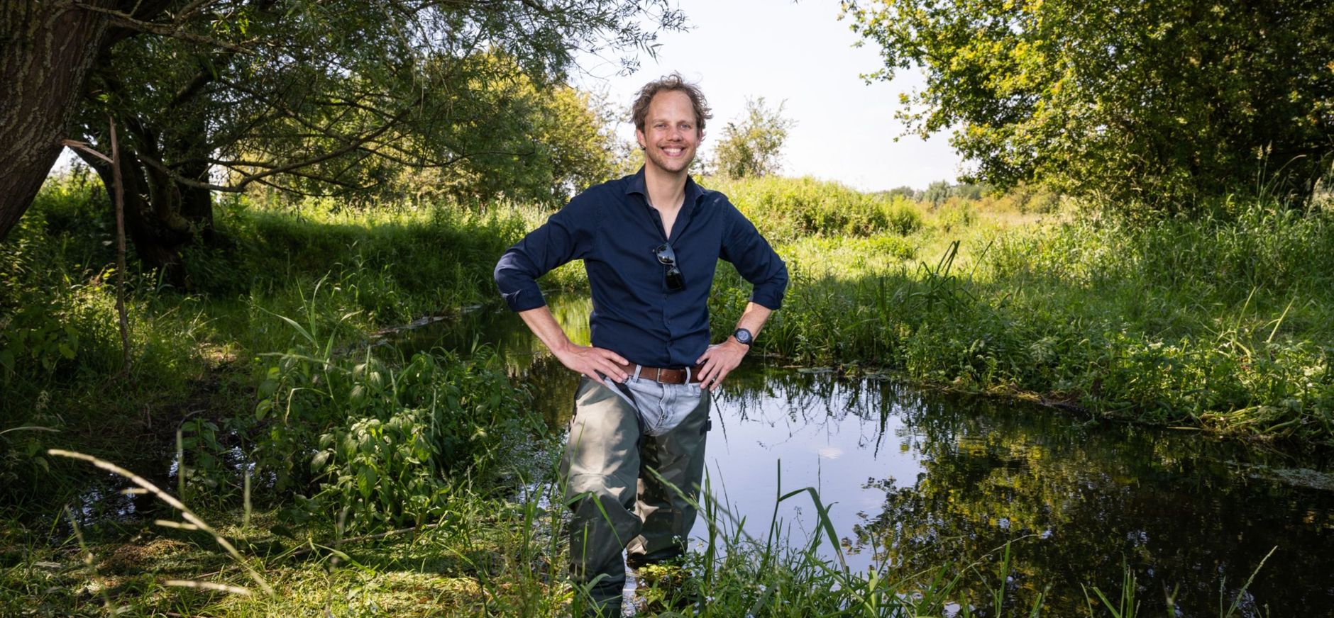 Man die tot net onder zijn middel in het water staat, hij heeft lieslaarsen aan en kijkt lachend in de camera
