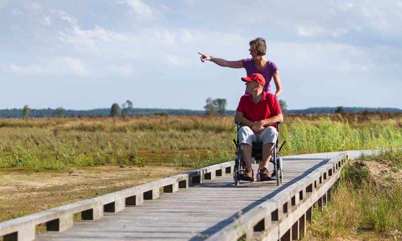 Vrouw loopt achter rolstoel waar een man in zit, over een houten bruggetje