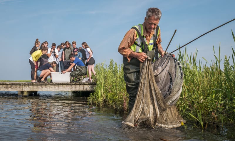 Man staat met groot visnet in zijn handen in het water, op de achtergrond kijkt een groep jongeren in een witte bak