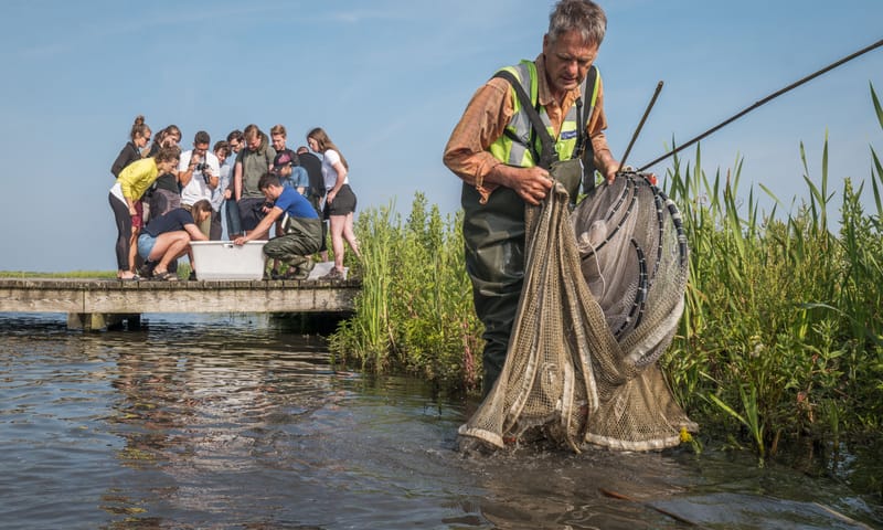 Man staat met groot visnet in zijn handen in het water, op de achtergrond kijkt een groep jongeren in een witte bak