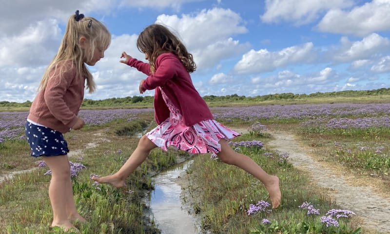 een meisje springt over een geultje op de kwelder. Een ander meisje staat op het punt ook te gaan springen.