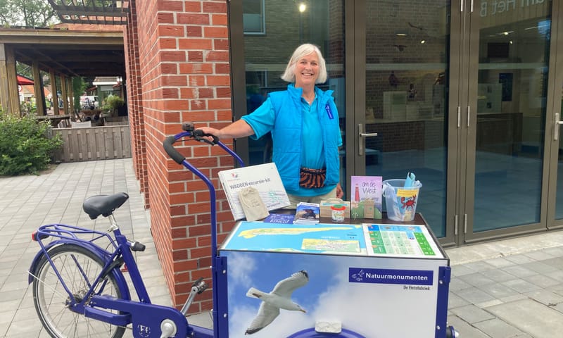 een vrolijke vrouw staat bij een bakfiets met info over natuurmonumenten