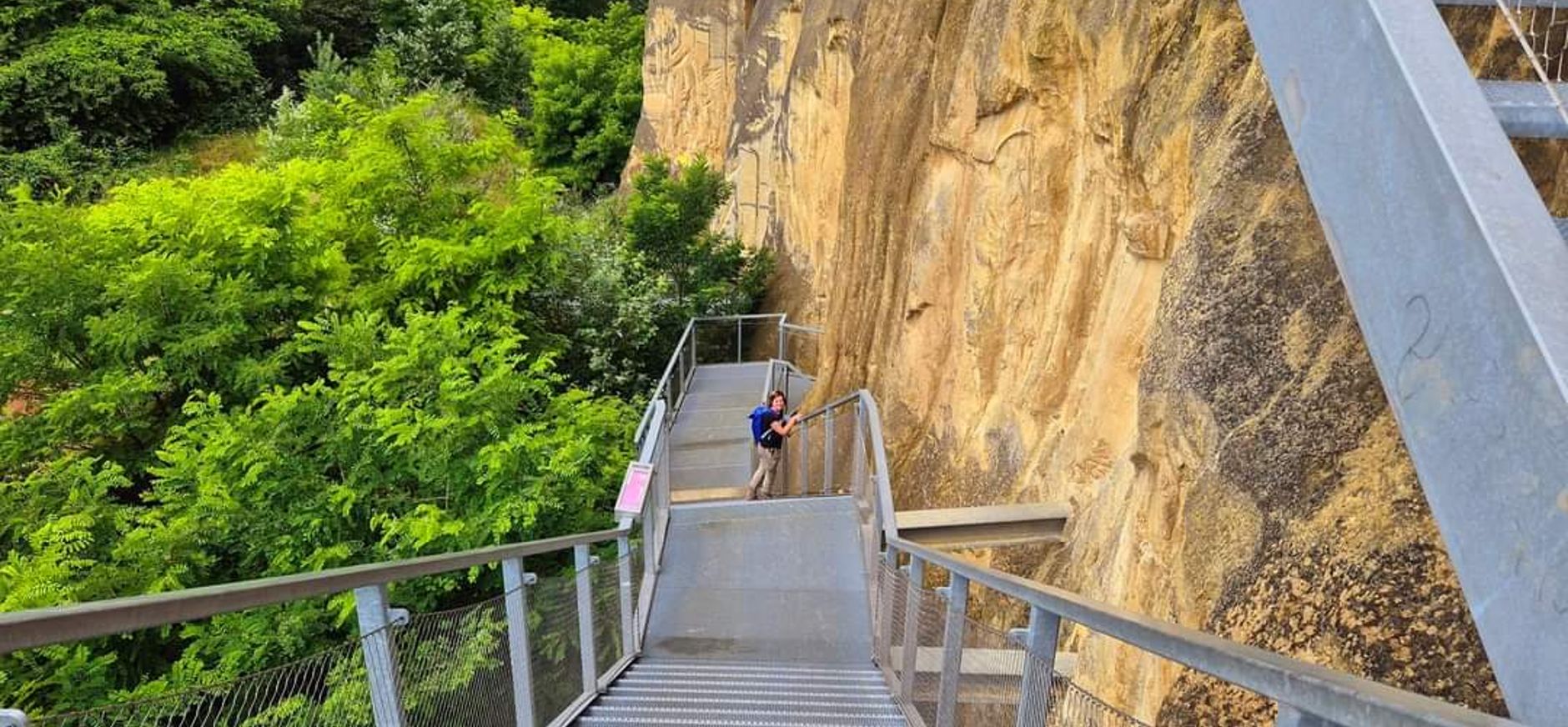 Foto van een vrolijke mevrouw die aan het wandelen is in natuurgebied Sint-Pietersberg. Ze staat stil en kijkt achterom tijdens het afdalen van de ijzeren trap die vanaf het plateau en het uitkijkplatform naar beneden leidt naar de ENCI-groeve. De foto is van bovenaf genomen. Je ziet links de diepte en groene begroeiing, in het midden de trap met daarop de mevrouw en rechts de steile kalkwand.