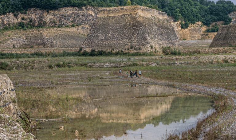 Gezinnen met kinderen wandelen met een gids tijdens excursie door de ENCI-groeve op de Sint-Pietersberg in Maastricht. Je ziet hoe groot de groeve is en hoe klein de mensen zijn. Op de foto zie je kalksteenwanden, waterpartijen, groene begroeiing en een klein beetje lucht.