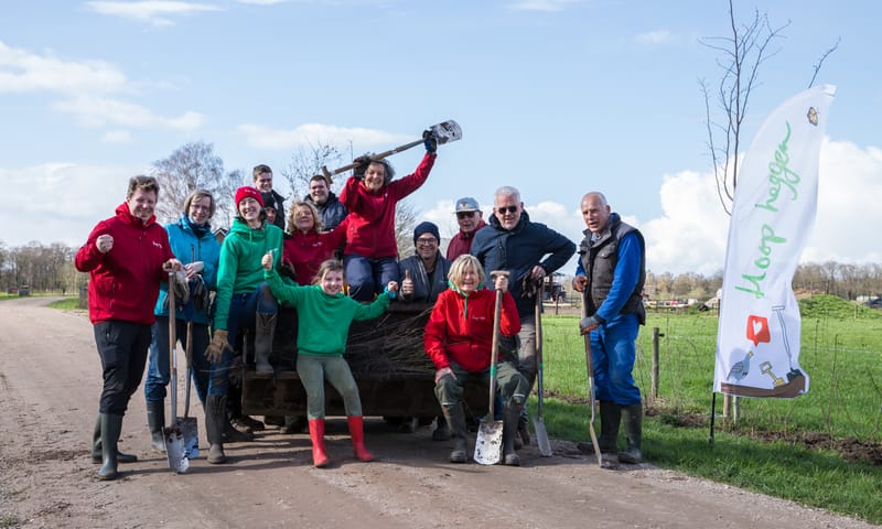 Groep mensen met spades en kaplaarzen, klaar om heggen te gaan planten
