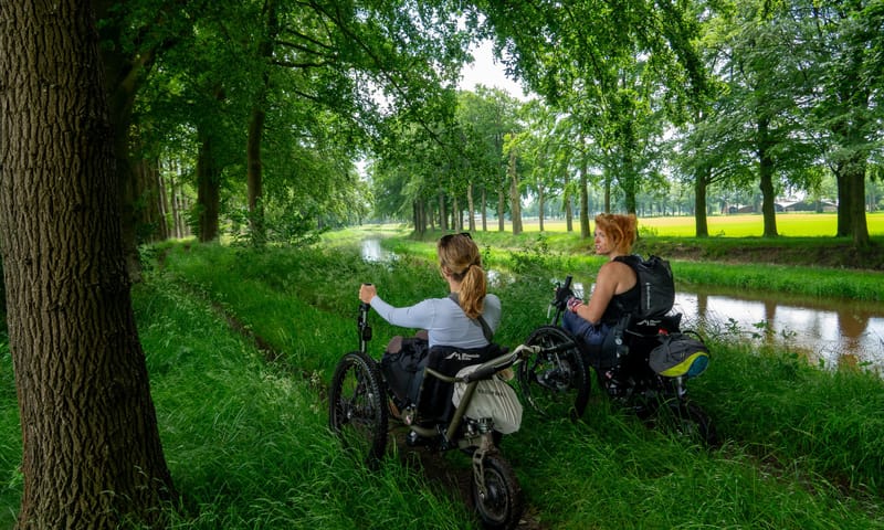 Twee dames in rolstoelen rijden over graspad door het bos