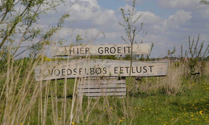 Houten bord tussen wat groen met de tekst 'hier groeit voedselbos eetlust'