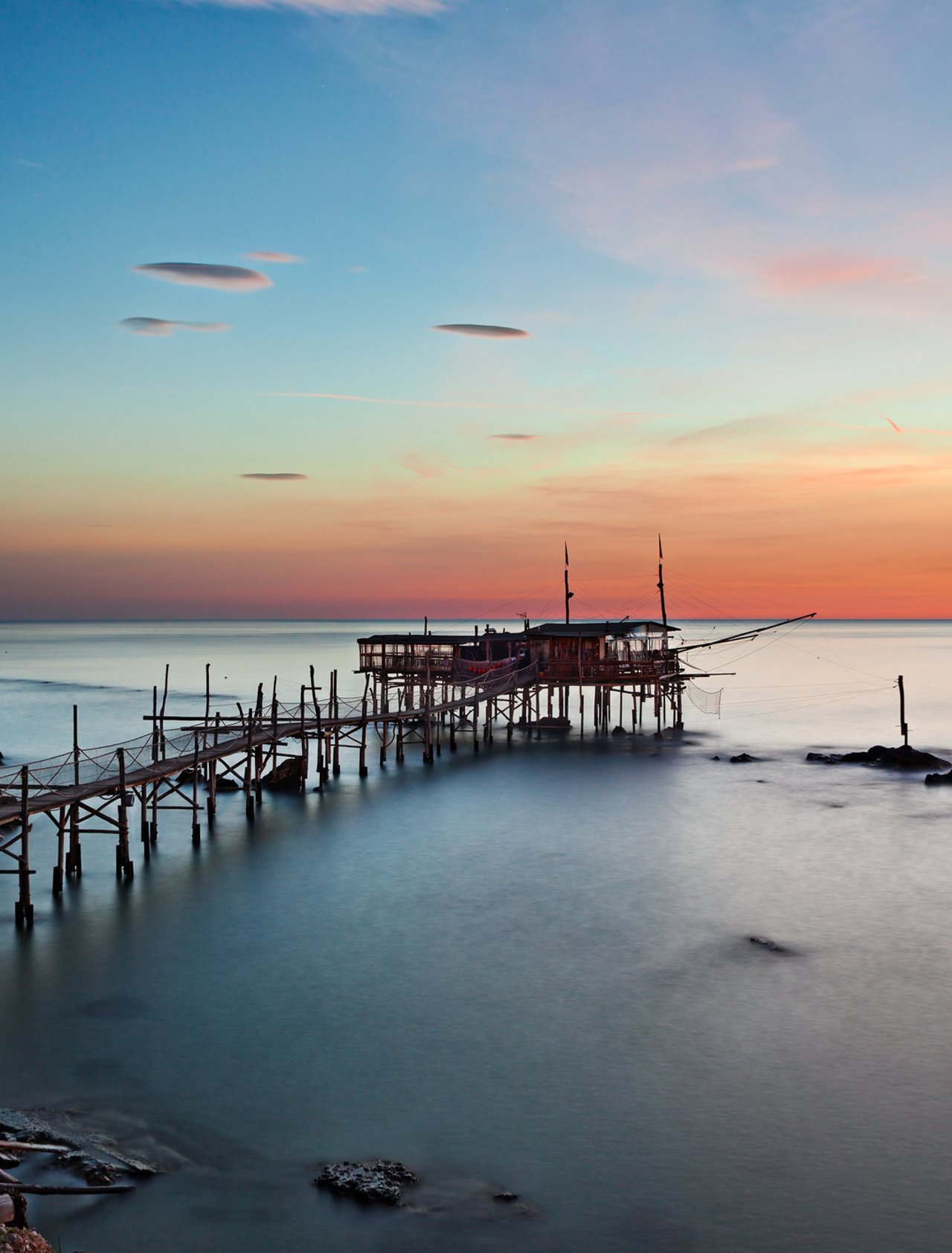 A trabocco at sunset in Chieti province, Abruzzo, with its long wooden walkway extending over still water.