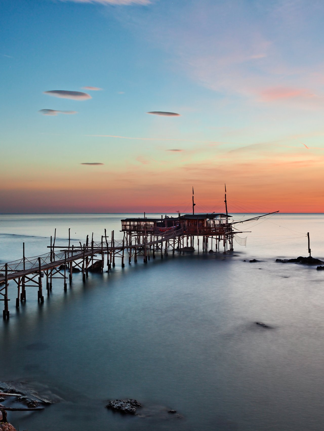 A trabocco at sunset in Chieti province, Abruzzo, with its long wooden walkway extending over still water.