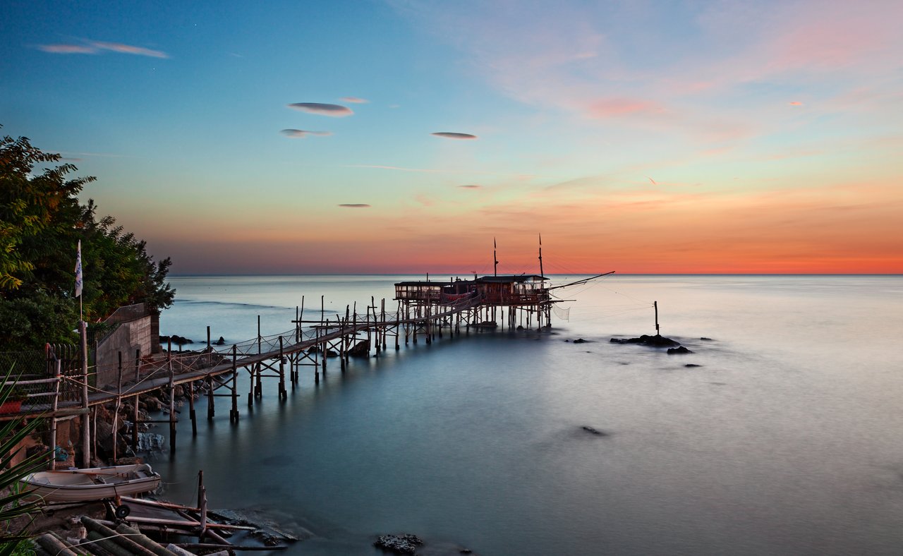 A trabocco at sunset in Chieti province, Abruzzo, with its long wooden walkway extending over still water.