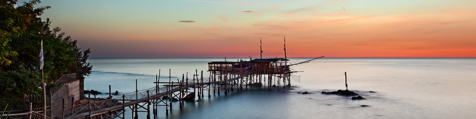 A trabocco at sunset in Chieti province, Abruzzo, with its long wooden walkway extending over still water.